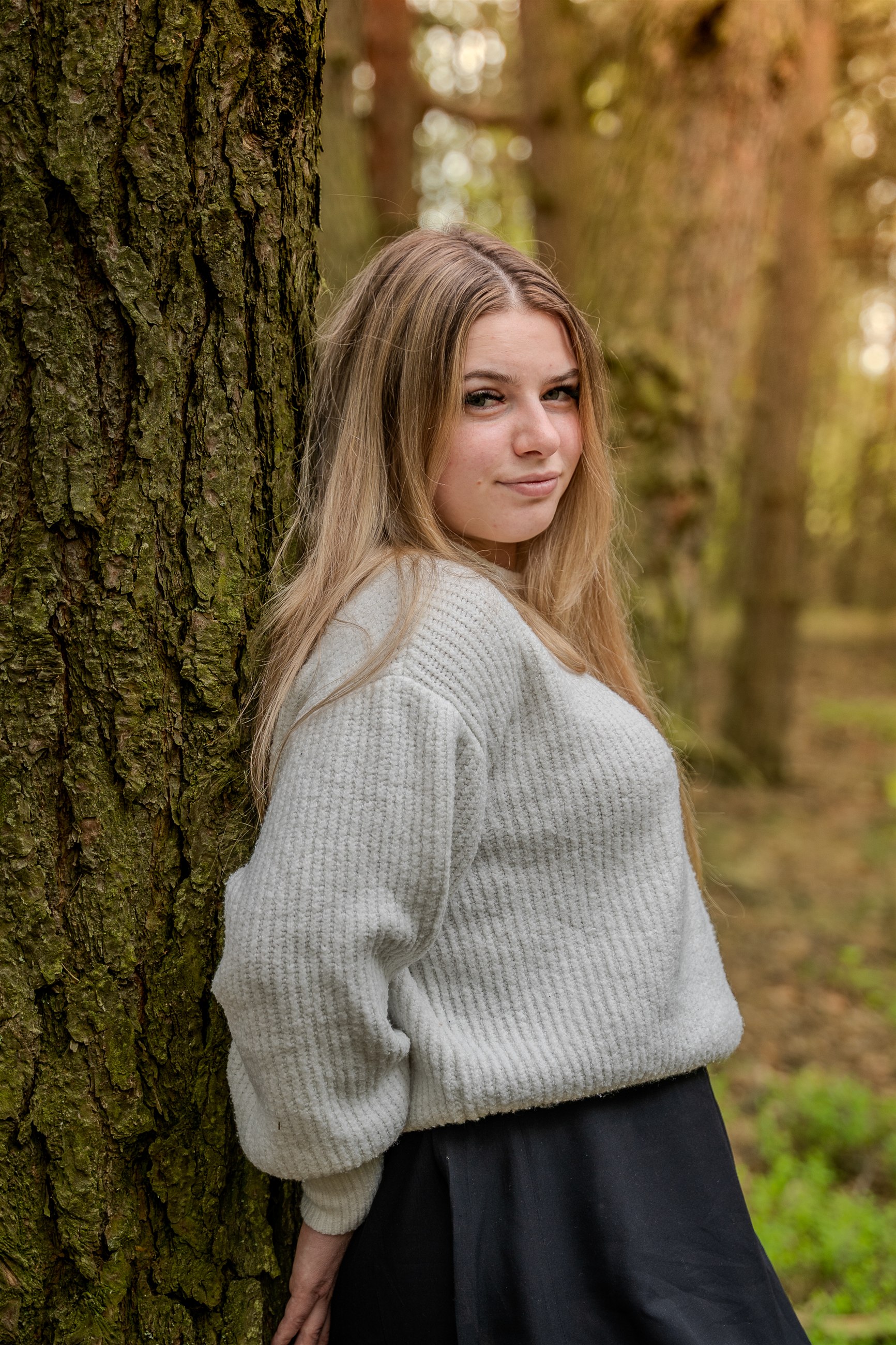 Naturalny portret kobiety w jasnym swetrze przy drzewie w lesie.
